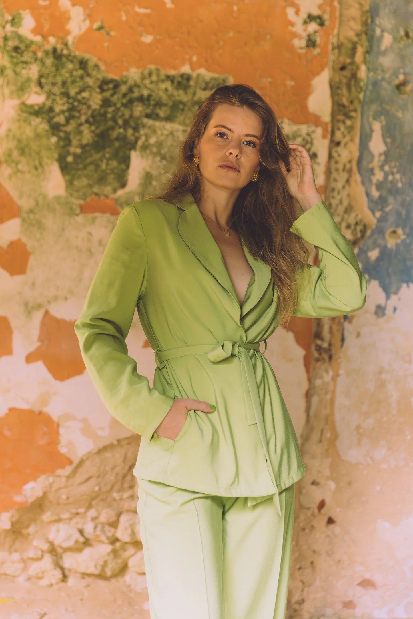 Woman wearing a light green belted blazer and matching pants standing against a rustic, textured wall background, showcasing the Siz blazer in a stylish and modern look.