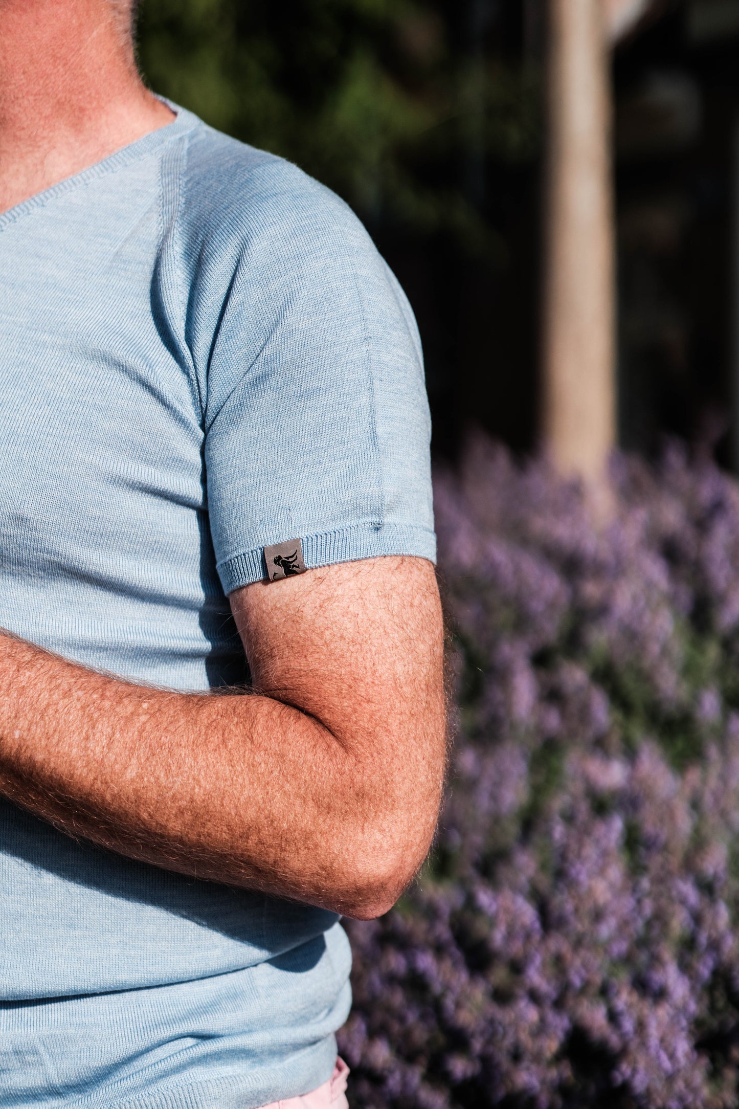 Primer plano del brazo de un hombre con camisa celeste de manga corta y una pequeña etiqueta con logo en la manga, mostrando la prenda Meedin con luz natural al aire libre.
