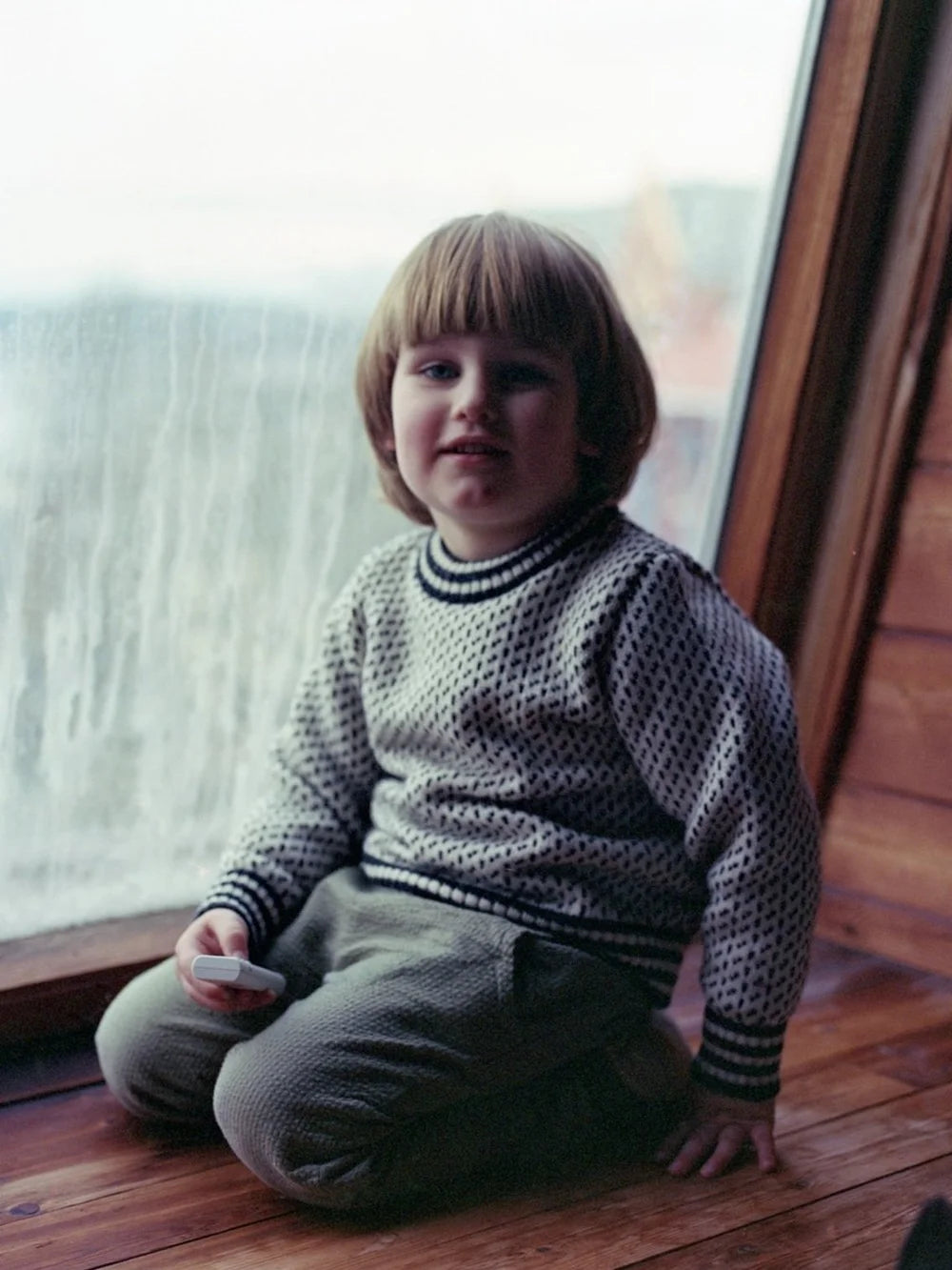 Niño sentado junto a una ventana neblinosa, con un suéter estampado y pantalones texturizados, mostrando la comodidad acogedora y cálida típica de Norlender Knitwear - Knitwear.