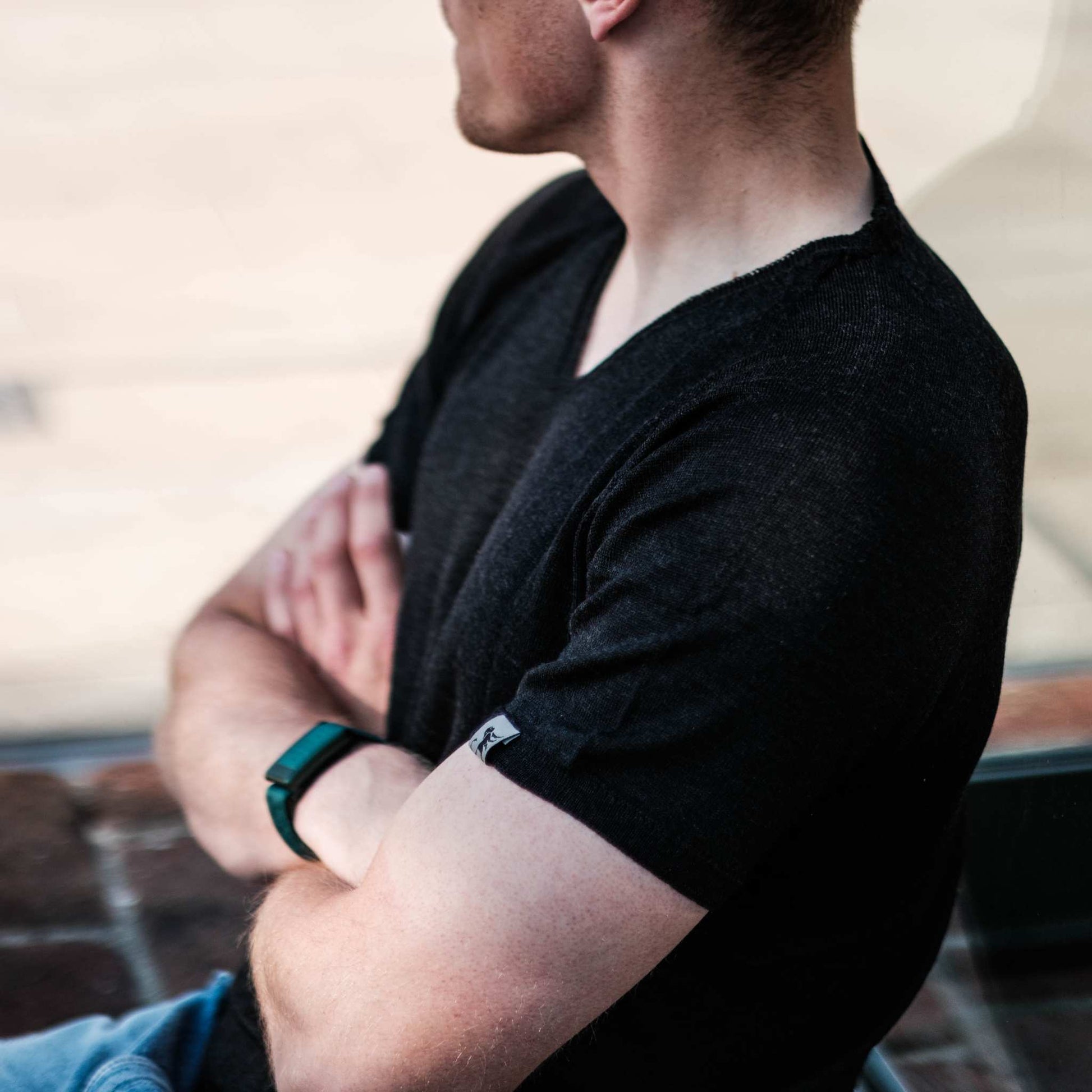 Hombre con camiseta negra de cuello en V con la marca Meedin, sentado con los brazos cruzados y mirando hacia un lado, con reloj de pulsera negro.