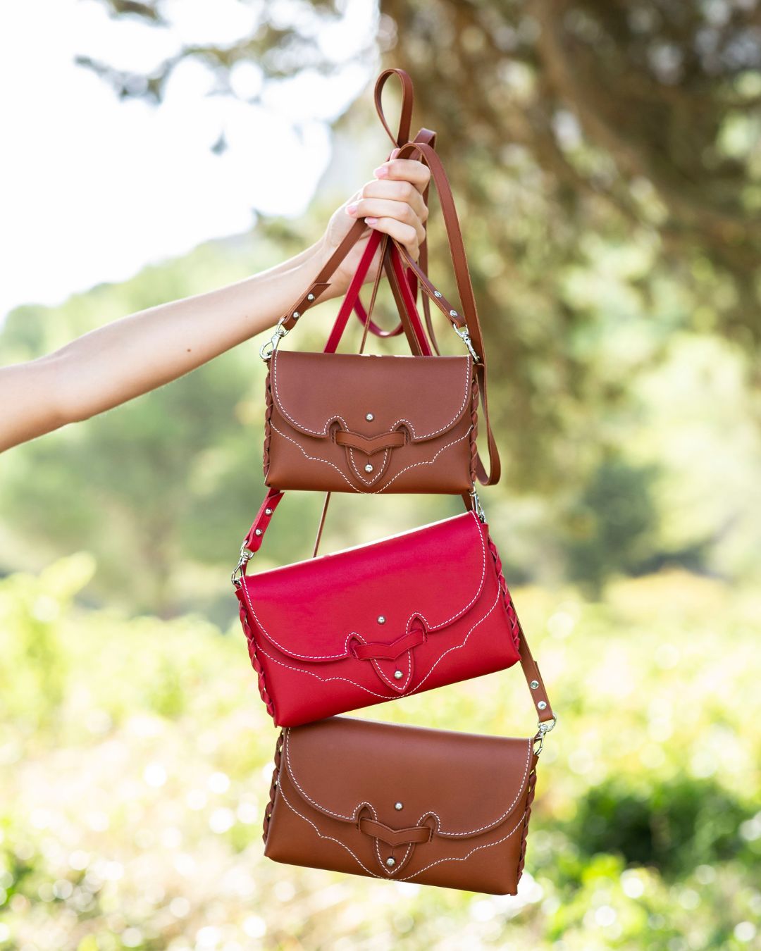 Three handbags in red and brown held against a blurred natural background