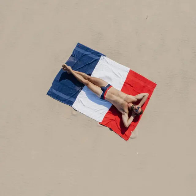 Hombre tumbado sobre una toalla con los colores de la bandera francesa en la arena, vistiendo Garçon Français - ropa interior con cinturilla azul y detalles rojos y blancos.