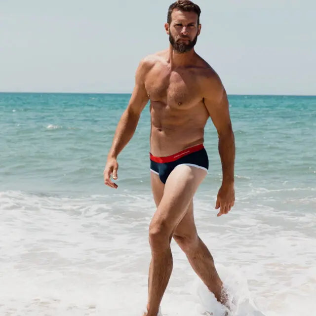 Hombre caminando por la playa con Garçon Français azul marino y rojo: calzoncillos tipo slip con olas del océano de fondo