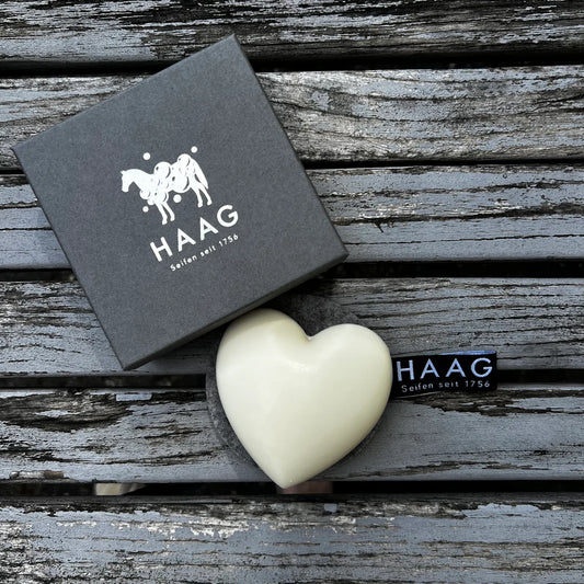 Zwarte verpakking met wit paardenlogo en tekst "HAAG Seifen seit 1756" naast een witte hartvormige zeep met zwart label, op een verweerd houten oppervlak, met Seifen Haag.