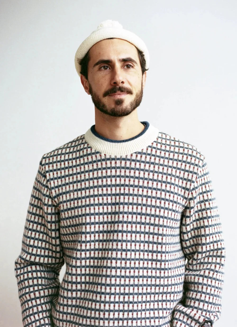 txt
Hombre con suéter de punto estampado con cuadros azules, blancos y rojos y gorro blanco, mostrando Norlender Knitwear, en un estilo casual.