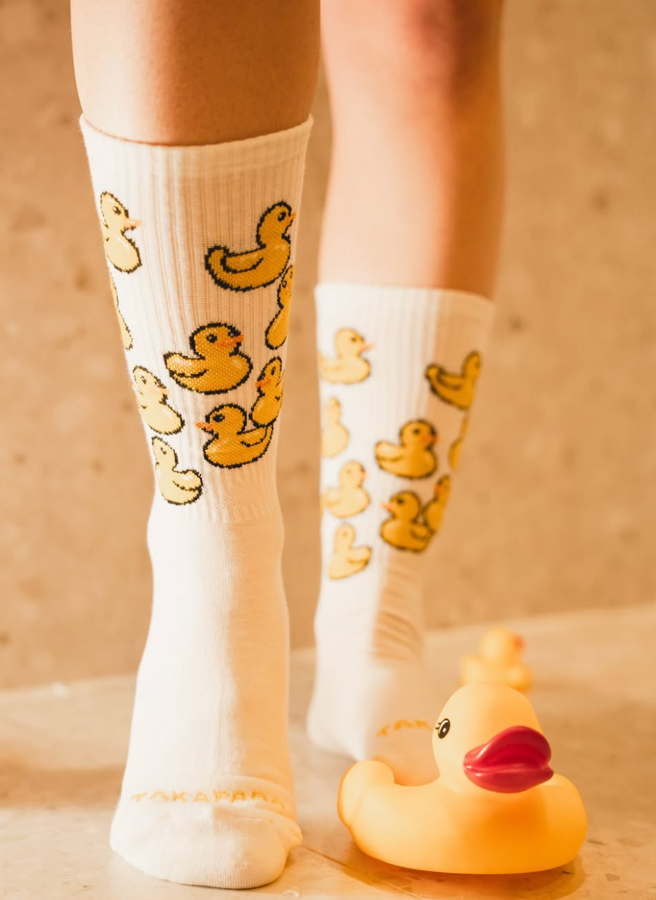 Persona con calcetines blancos con múltiples patrones de patitos de goma amarillos y el texto Takapara - Socks en los dedos de los pies, de pie junto a un pato de goma sobre un suelo beige.