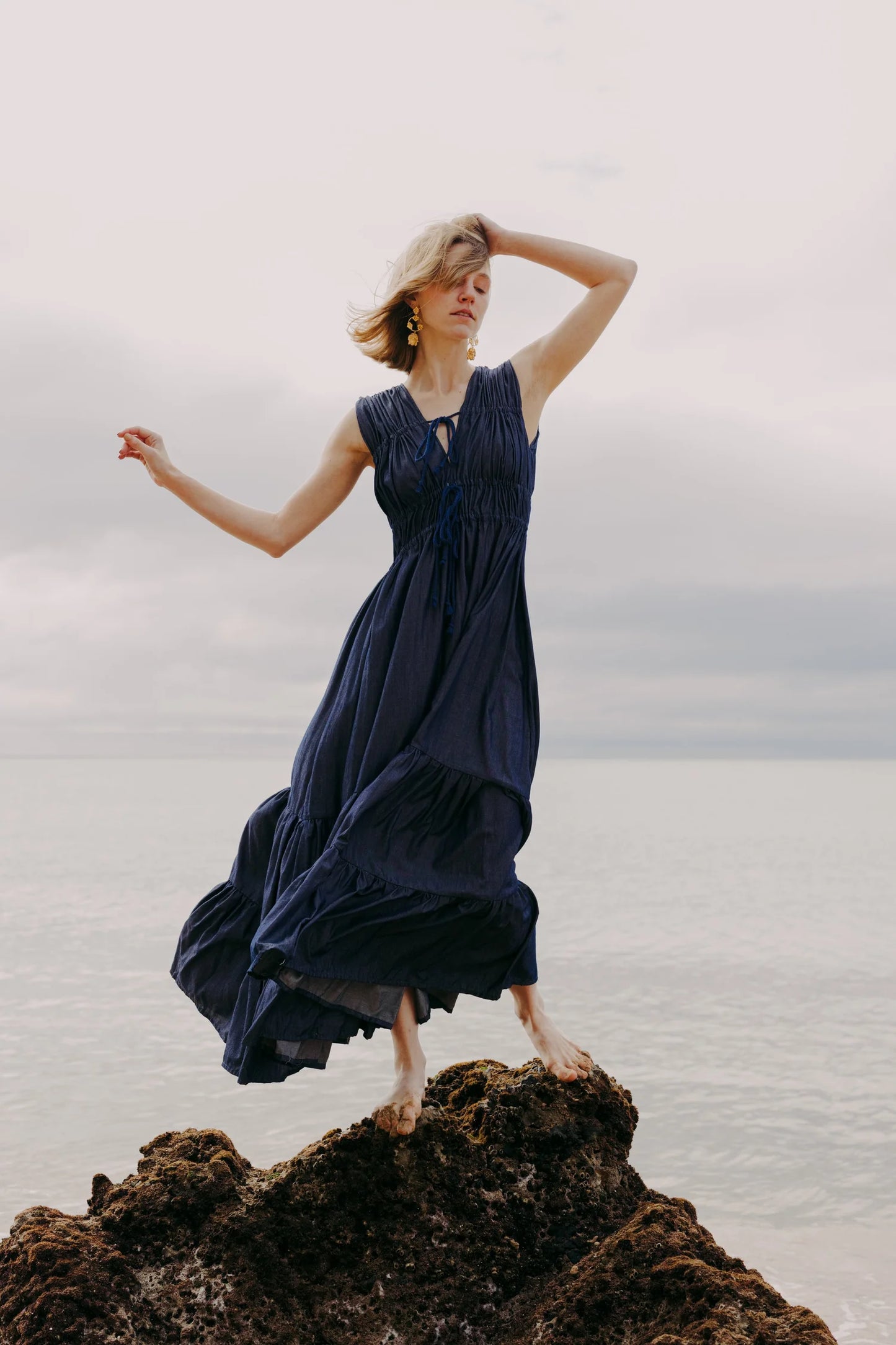 Woman wearing a flowing navy blue Siz dress standing barefoot on a rocky shore with the ocean and cloudy sky in the background, posing with one arm raised above her head.