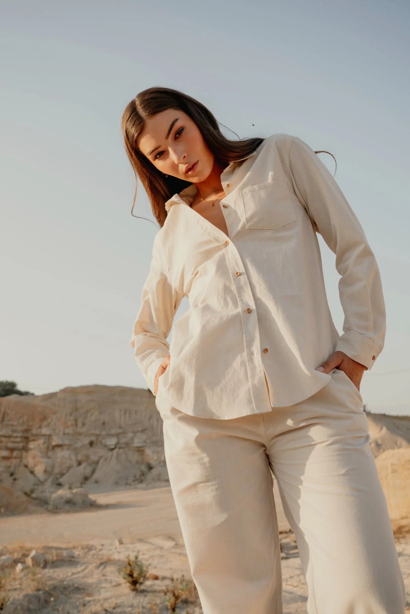 Woman wearing a light beige button-up shirt and matching pants outdoors, modeling the Siz shirt with a relaxed fit and casual style in a natural setting.
