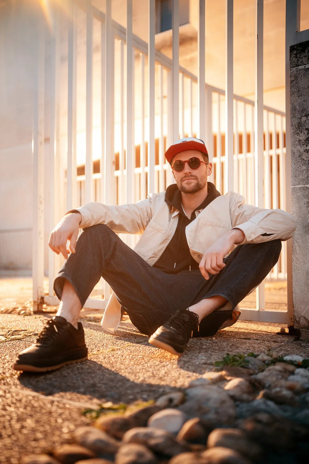 Hombre con gafas de sol, gorra roja, chaqueta ligera y zapatos negros sentado relajadamente en el suelo junto a una valla blanca durante la hora dorada, mostrando el estilo Ango.