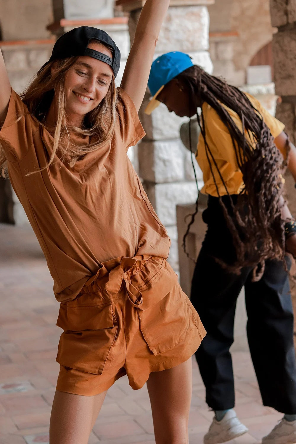 Young woman wearing a casual rust-colored Ango outfit consisting of a t-shirt and shorts, smiling and dancing with arms raised, accompanied by another person in casual attire.