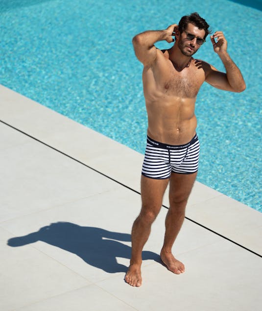 Un hombre en forma con ropa interior Garçon Français a rayas está descalzo junto a una piscina azul brillante, ajustándose las gafas de sol en un día soleado.