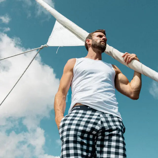 Hombre con camiseta blanca sin mangas y pantalones a cuadros de pie al aire libre junto al mástil de un velero bajo un cielo azul con nubes, mostrando Garçon Français - ropa interior.