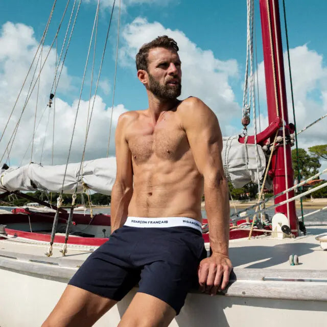 Man wearing navy blue Garçon Français - Underwear shorts sitting on a boat with sails and rigging in the background under a partly cloudy sky.